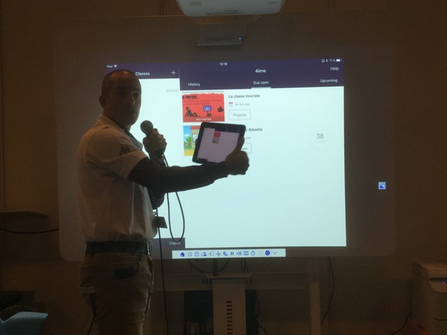 Le présentateur montre sur une tablette l'interface, aussi visible sur un écran numérique derrière lui.